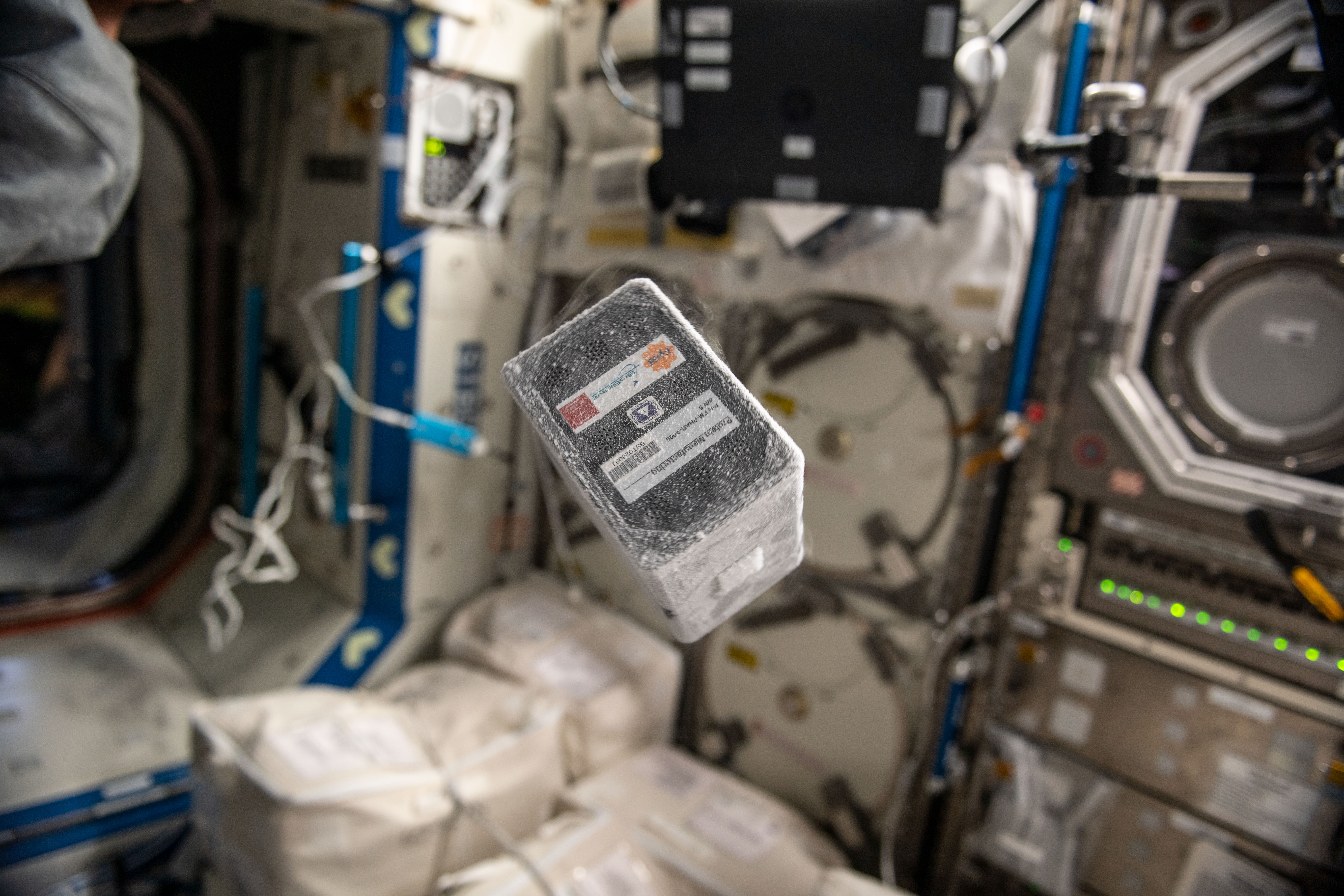 A frozen eight inch by four inch by six inch experiment floats aboard the International Space Staion, with equipment in the background.